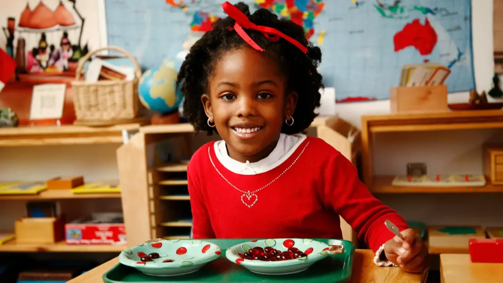 montessori-student-red-bead-materials Smiling girl using tweezers to move red counters on green plates in class.