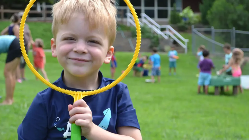 Young boy smiling through a yellow bubble-wand ring on a grassy field.