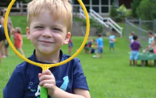 Young boy smiling through a yellow bubble-wand ring on a grassy field.