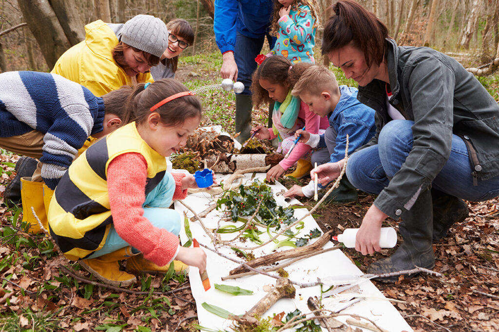 Children and teachers kneel on the forest floor, examining leaves, logs, and soil as part of a hands-on outdoor science exploration.