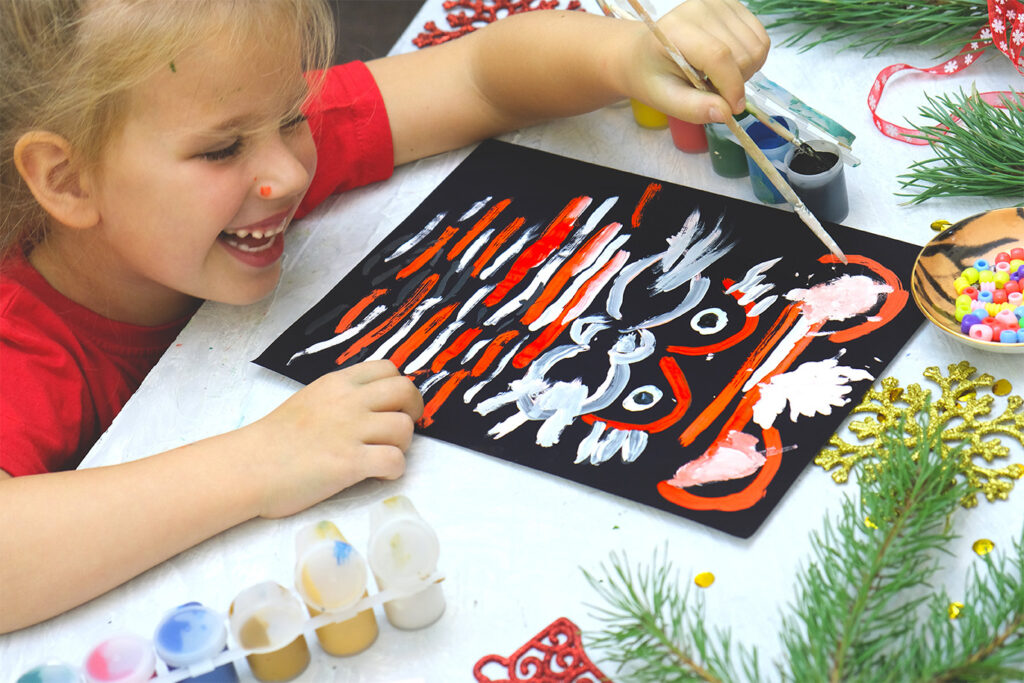 A young child joyfully painting a festive holiday design during a Montessori-inspired Christmas activity, surrounded by art materials and evergreen decorations.