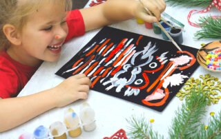 A young child joyfully painting a festive holiday design during a Montessori-inspired Christmas activity, surrounded by art materials and evergreen decorations.